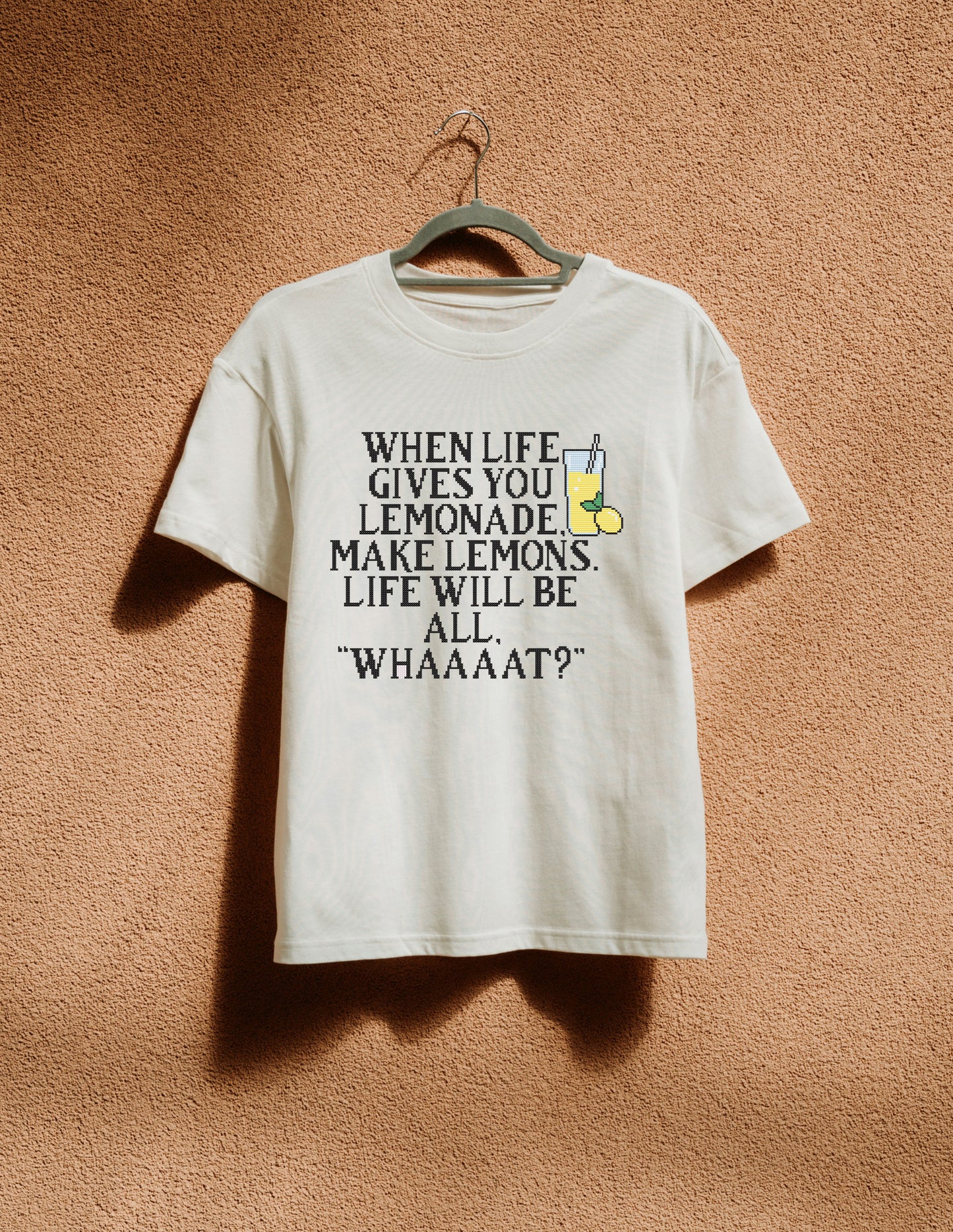 White t-shirt with humorous text design on a hanger against a textured brown wall.
