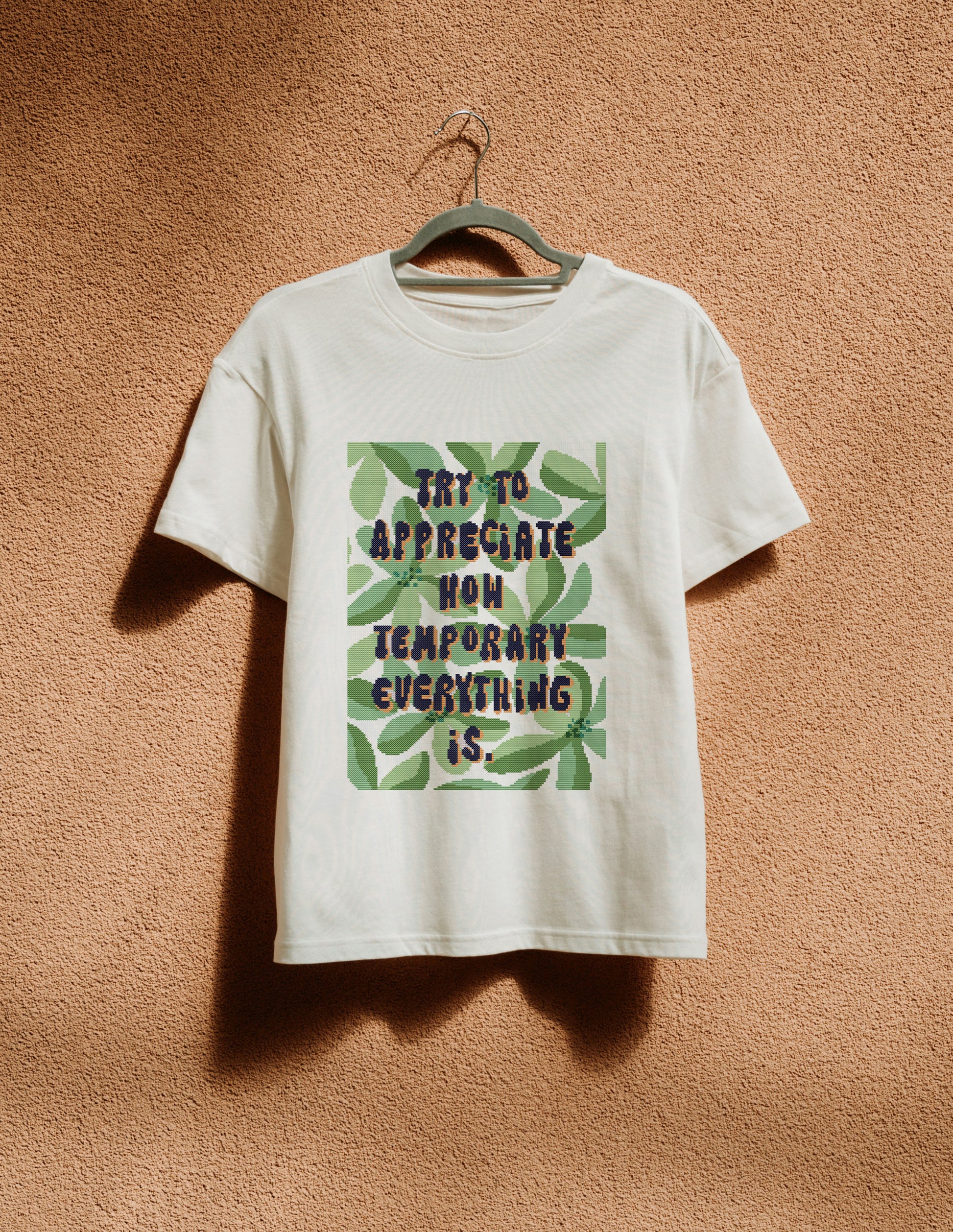 White t-shirt with text and leaf design on a hanger against a textured brown wall