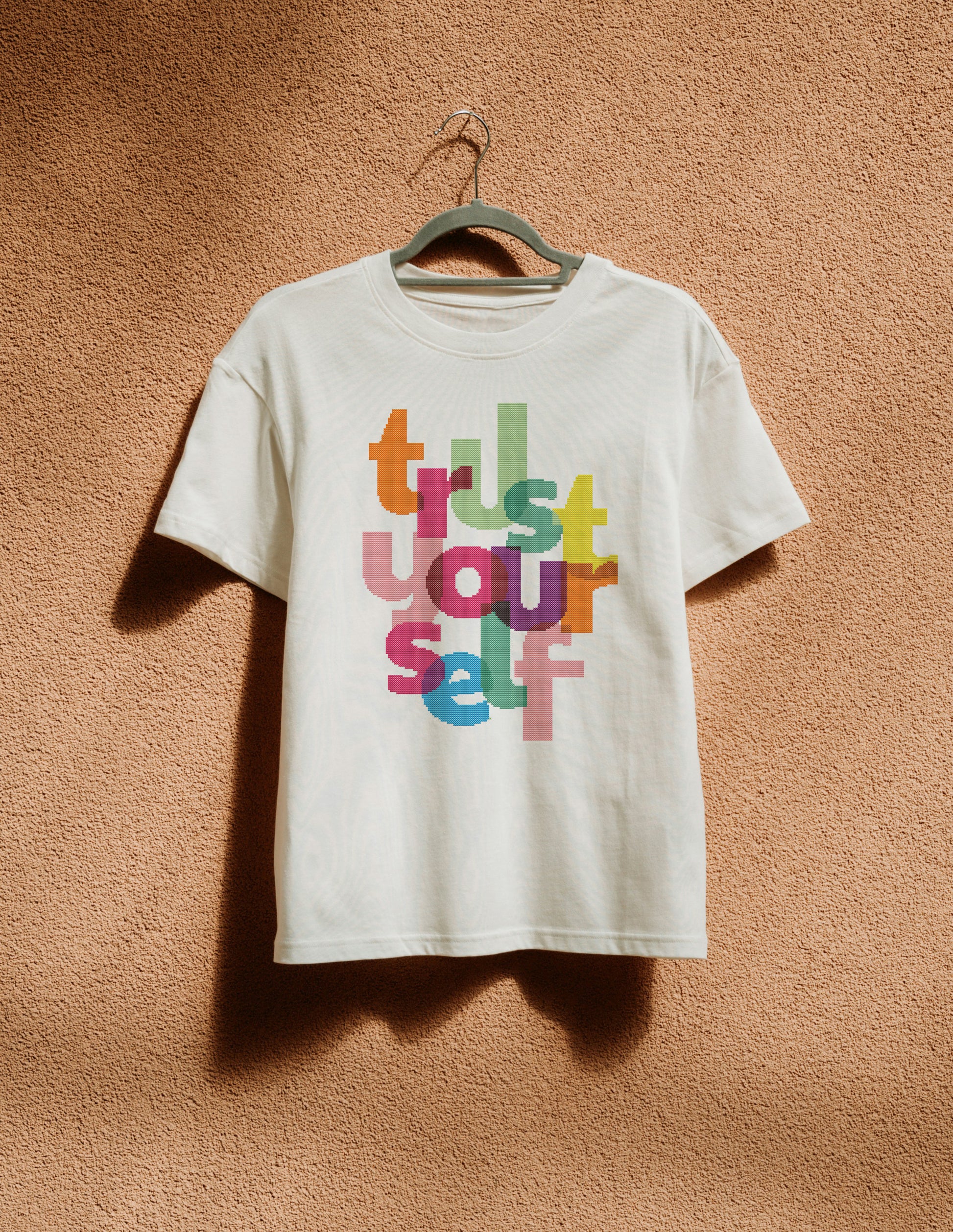 White t-shirt with colorful text 'Trust Yourself' on a hanger against a textured brown wall.