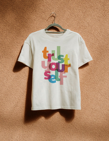 White t-shirt with colorful text 'Trust Yourself' on a hanger against a textured brown wall.