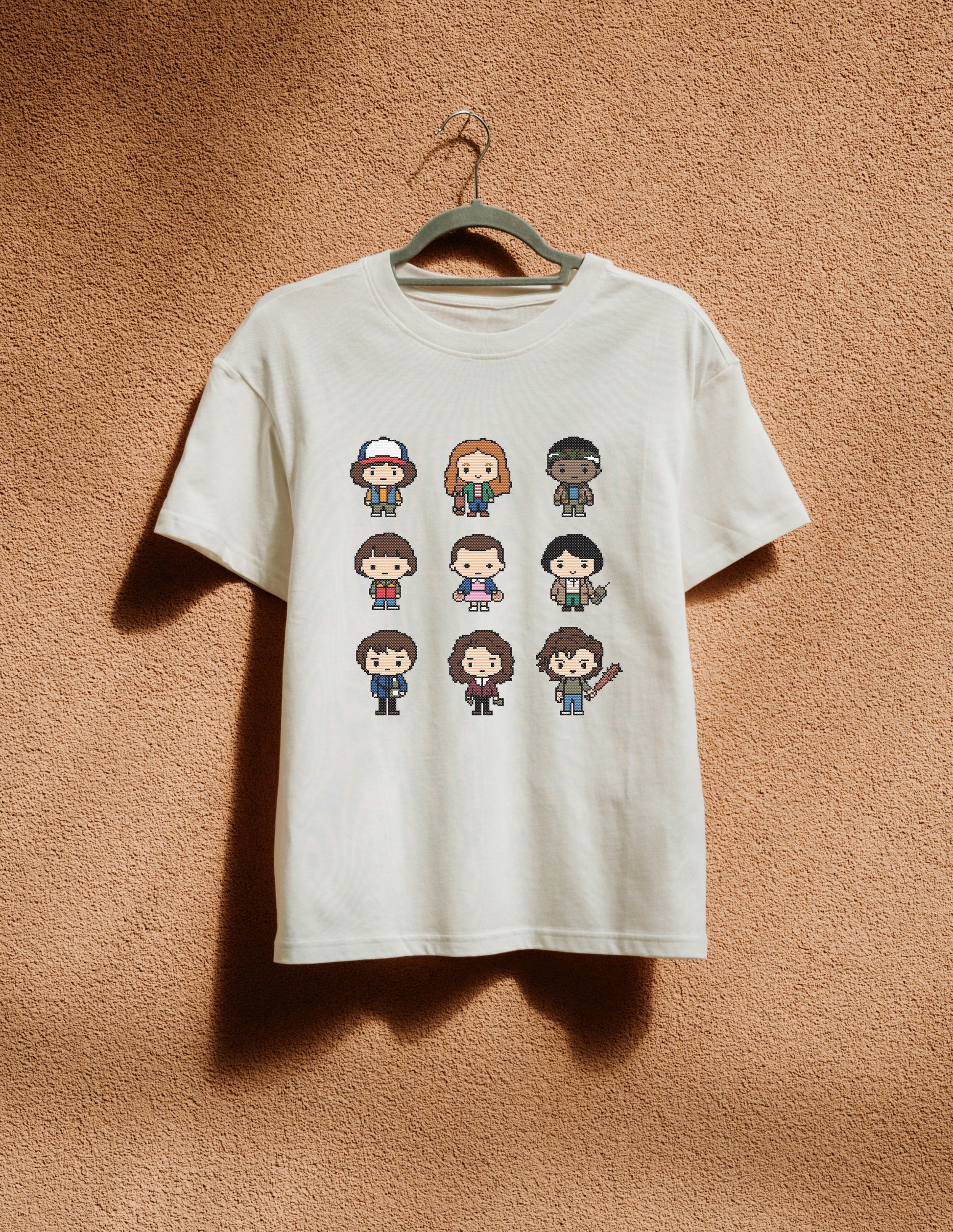 White t-shirt with pixelated character designs on a hanger against a brown textured wall.