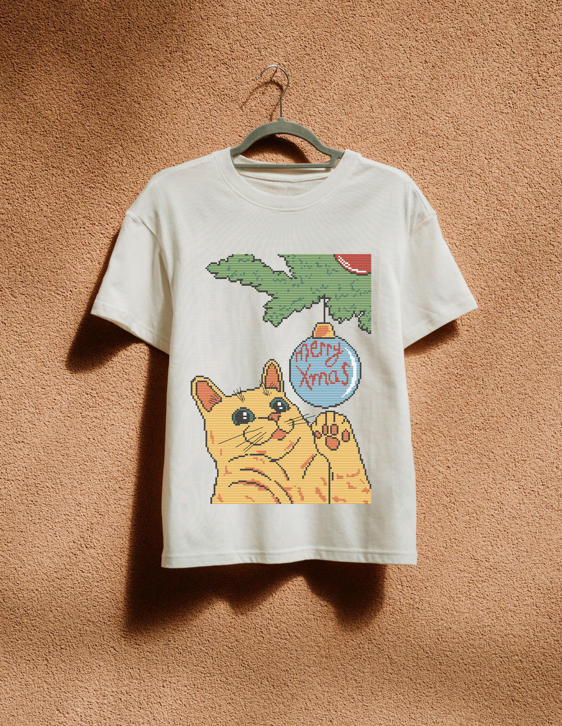White t-shirt with a cat holding a 'Merry Xmas' ornament graphic on a brown background