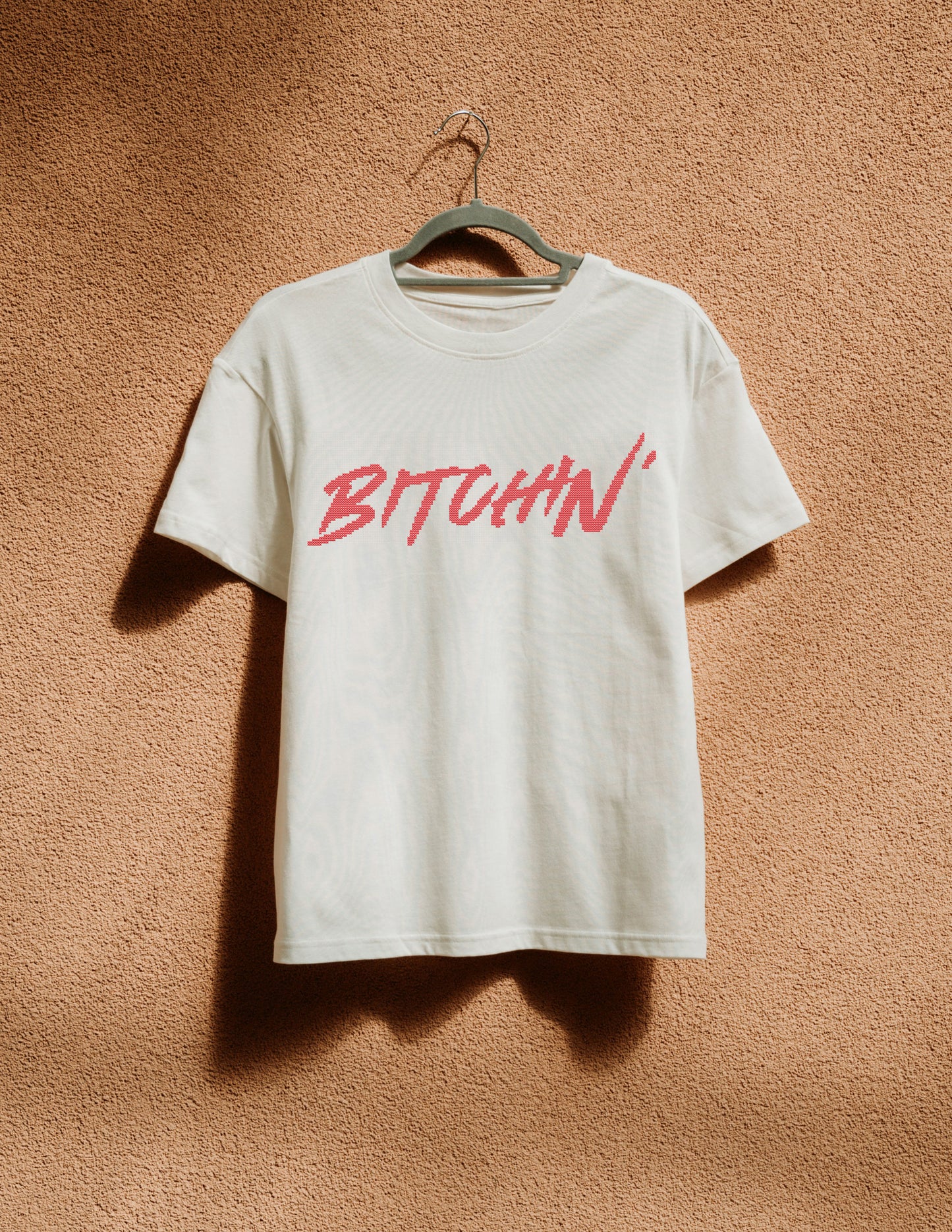 White t-shirt with 'BITCHIN'' in red on a hanger against a brown textured wall.