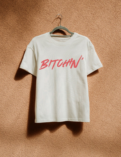 White t-shirt with 'BITCHIN'' in red on a hanger against a brown textured wall.