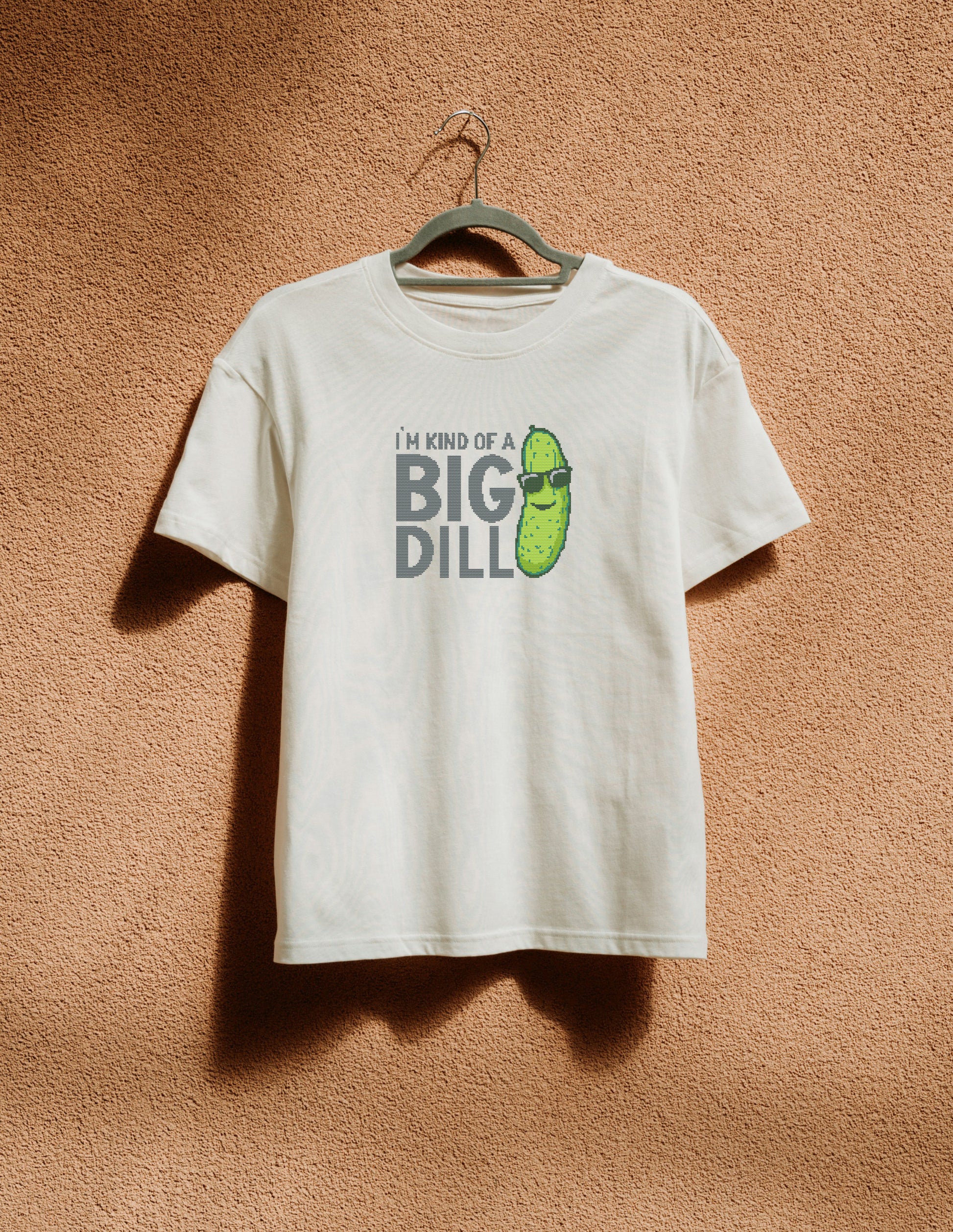 White t-shirt with pickle graphic and text on a hanger against a brown textured wall